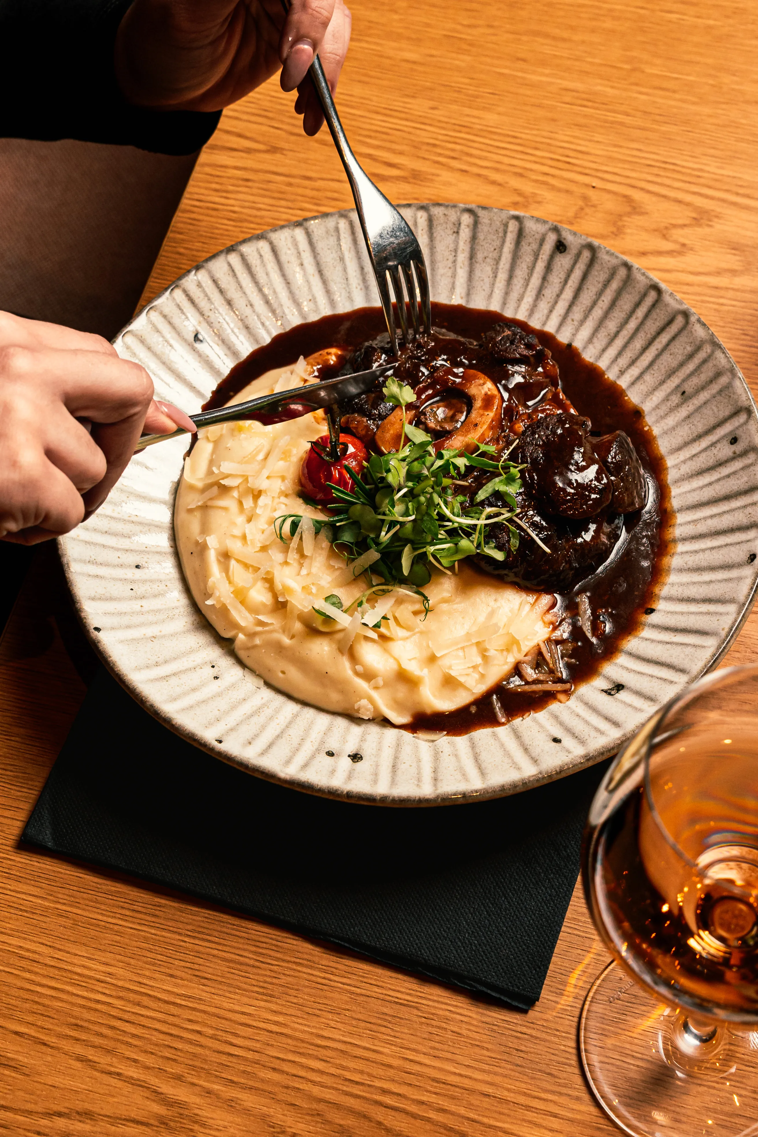 Prato de purê de batatas servido com carne e molho, decorado com ervas frescas e pimentas, sendo cortado com garfo e faca por uma pessoa; taça de vinho ao lado sobre uma mesa de madeira.